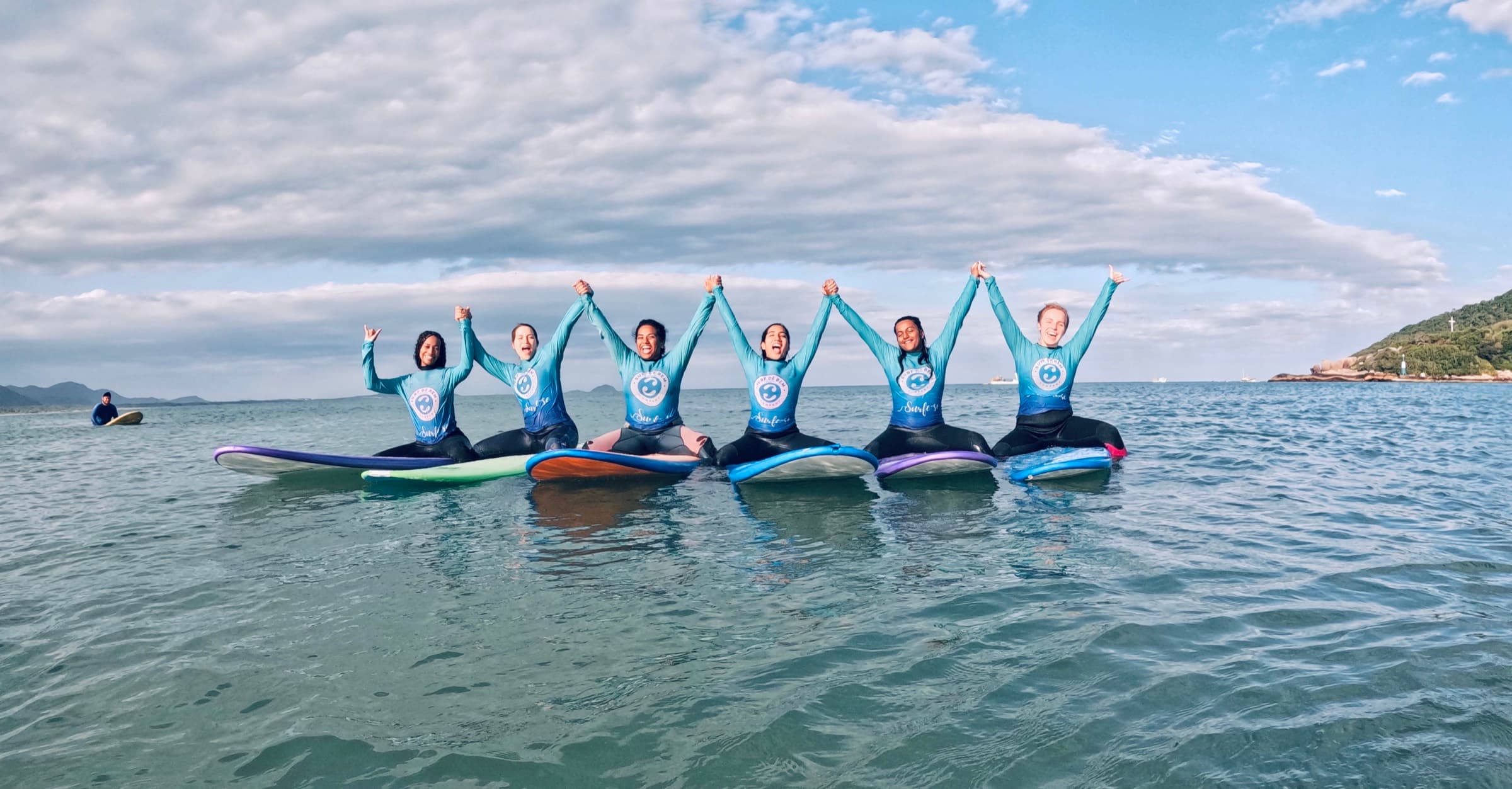 Grupo de surfistas em suas pranchas na água, de mãos dadas e braços para o alto.